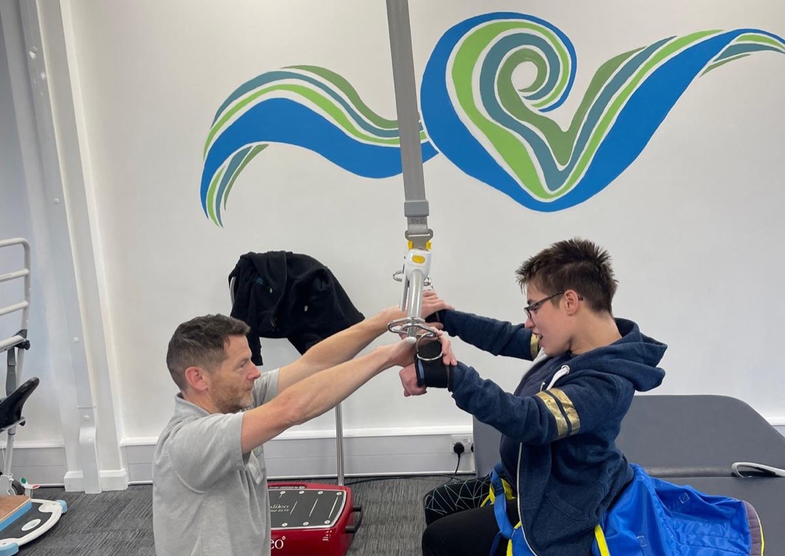 A physiotherapist working with a client using the ceiling hoist in the Get Ahead clinic gym, beneath the clinic's branded wall mural