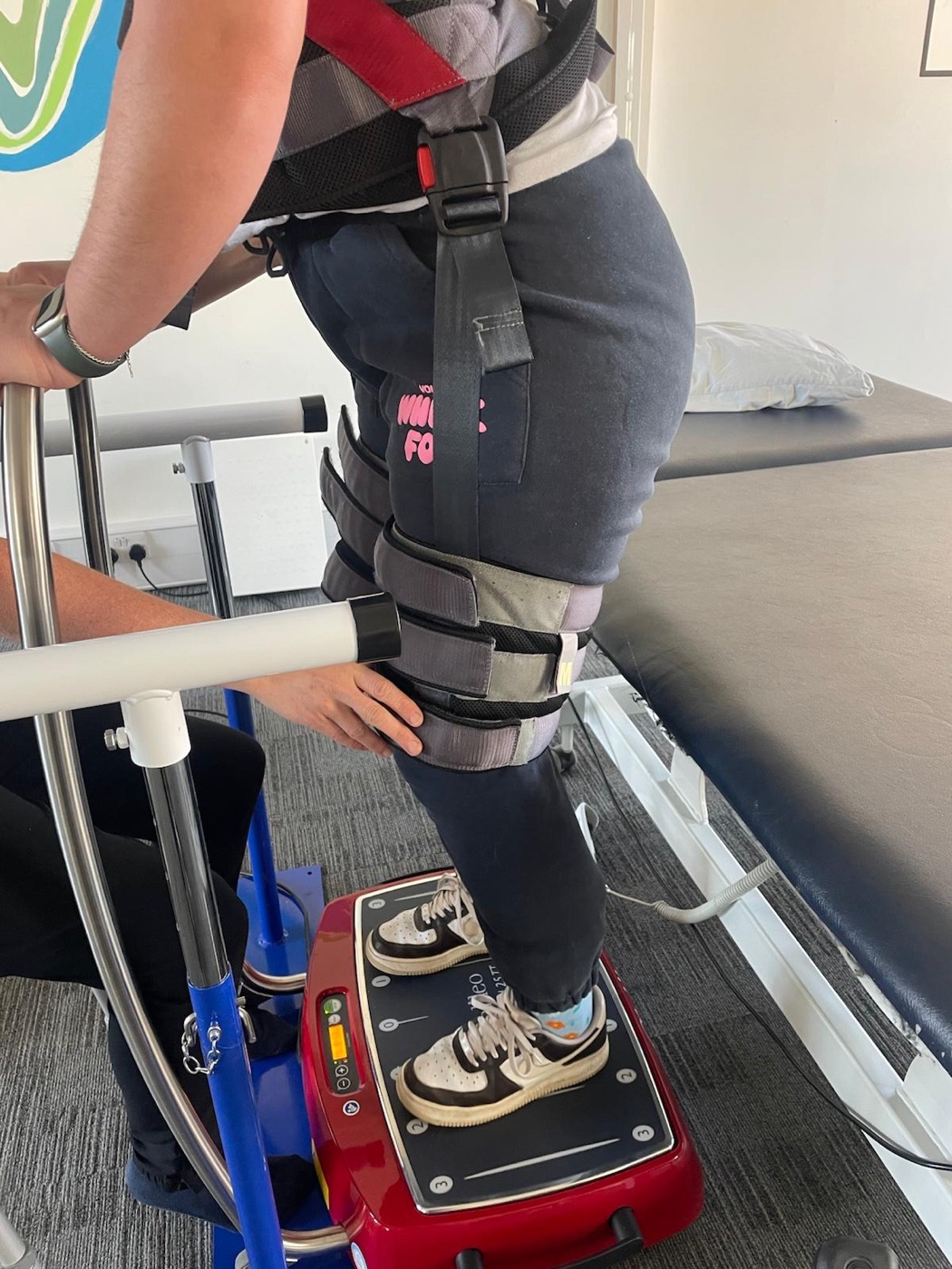 An adult standing on the Galileo vibration plate in a support frame while a physiotherapist steadies their knee